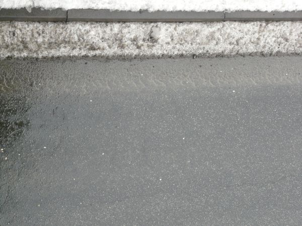 Road edge texture, with grey asphalt embedded with white rocks meeting edges of black-flecked snow, grey sidewalk and a white snow with grey patches.